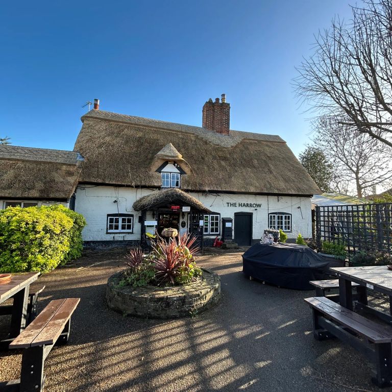 A traditional white pub with a thatched roof, outdoor seating, and planters under a sunny blue sky.