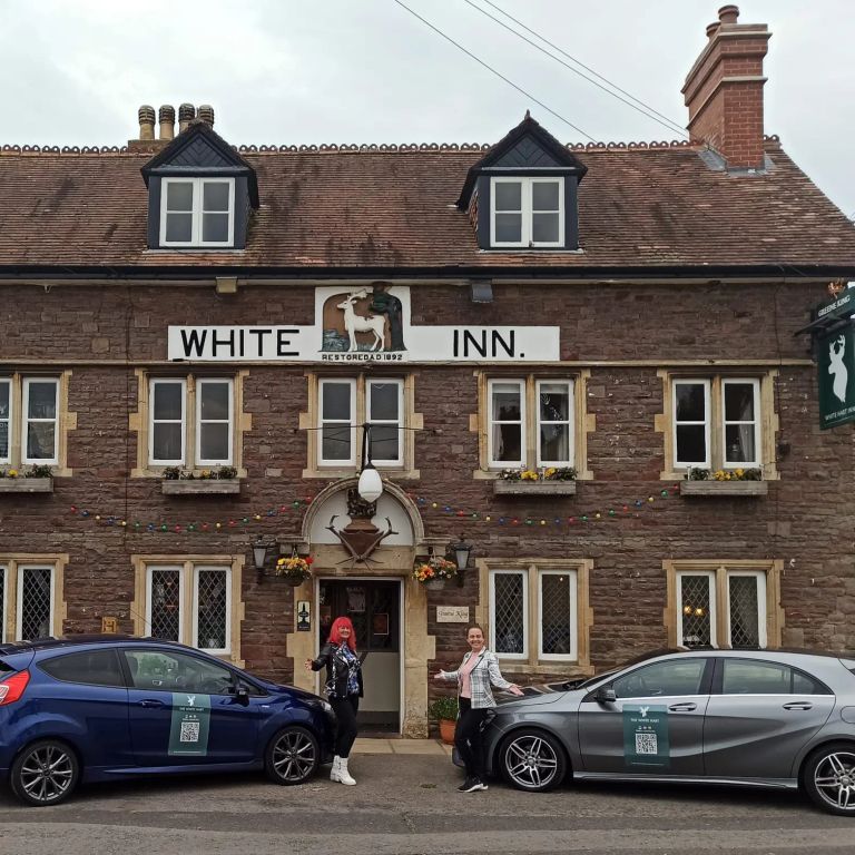 Two people stand outside the historic brick White Inn, with a blue and a silver branded car parked in front.