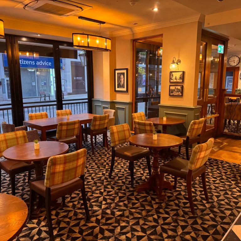 Pub interior with plaid chairs, geometric floor, and tables near large street-facing windows.