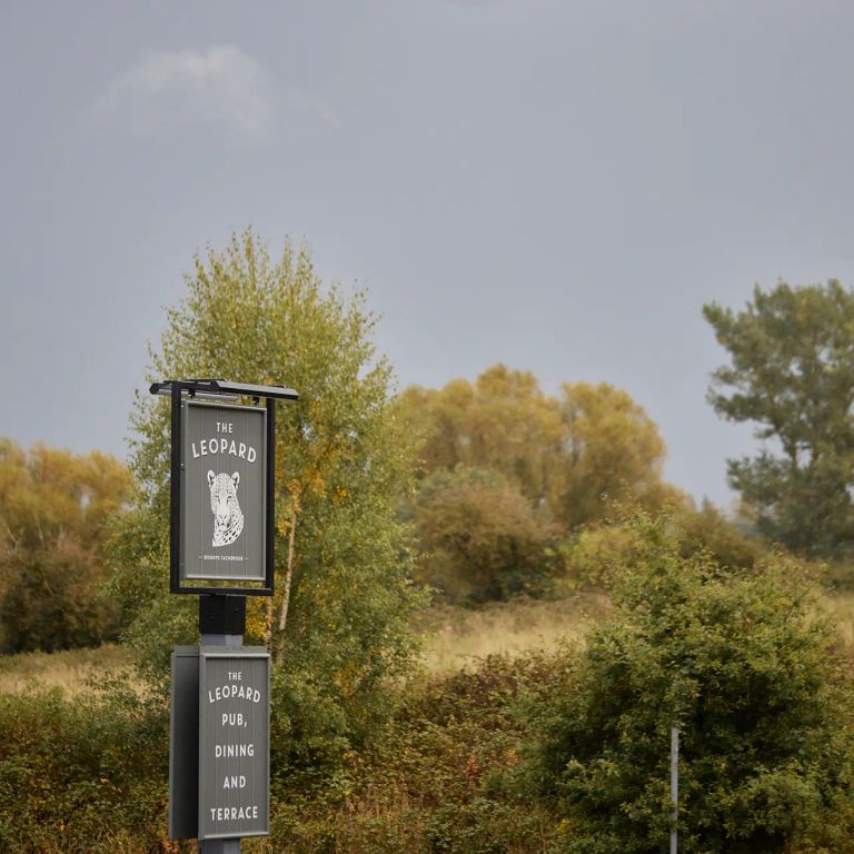 Pub sign for "The Leopard" with a leopard logo, set among trees and bushes under an overcast sky.