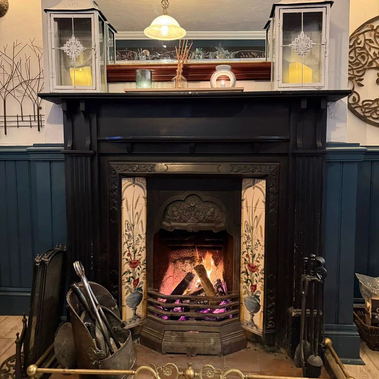 Ornate fireplace with a burning fire, decorative tiles, a dark mantel, two lanterns, and dark blue wainscoting.